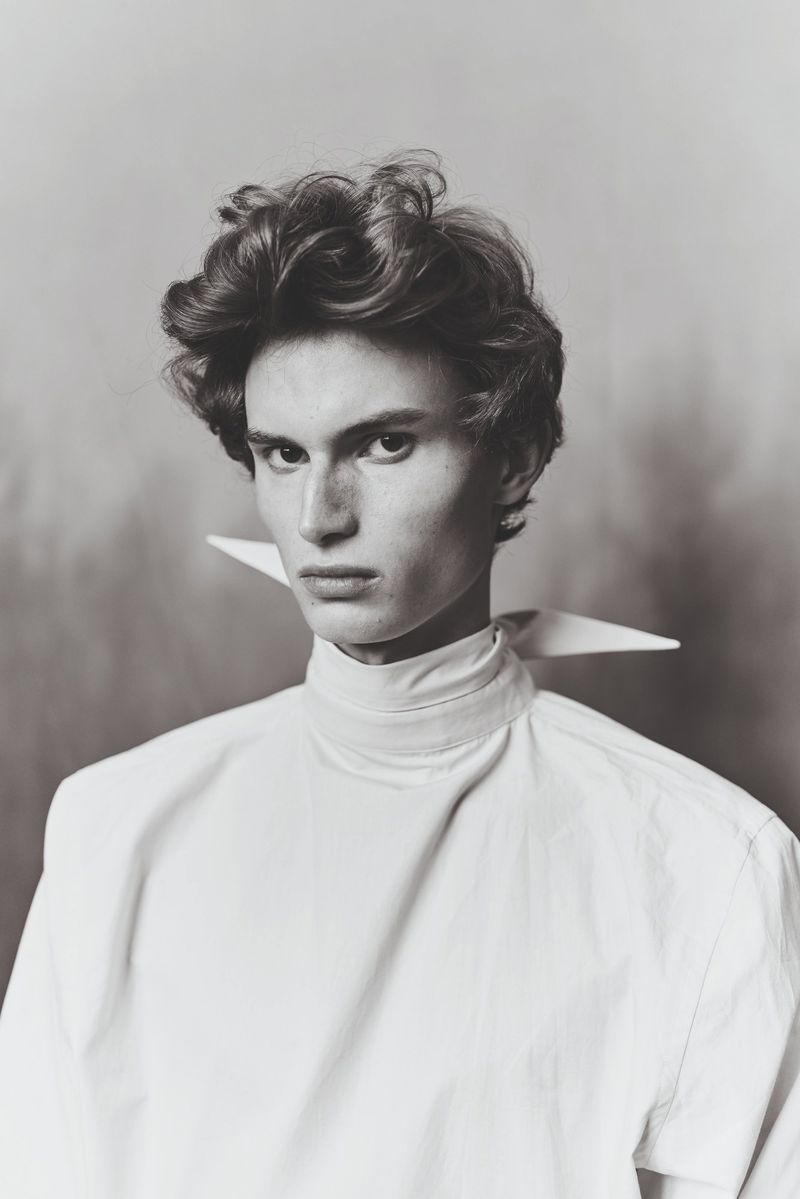 Monochromatic portrait of a young adult male with wavy hair wearing a white, high-collared shirt against a blurred background.