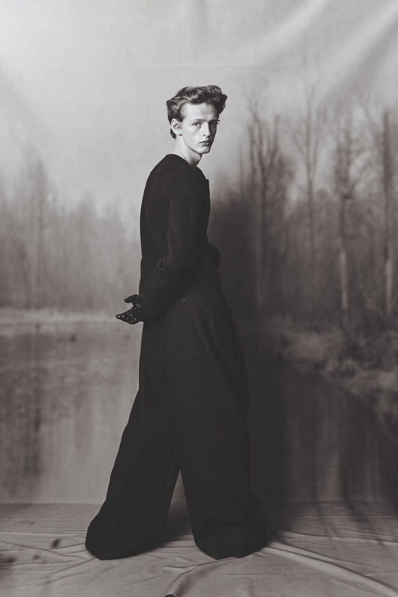 Fashion portrait of a young adult male in dark clothing against a blurred nature backdrop, captured with artistic monochromatic tones.