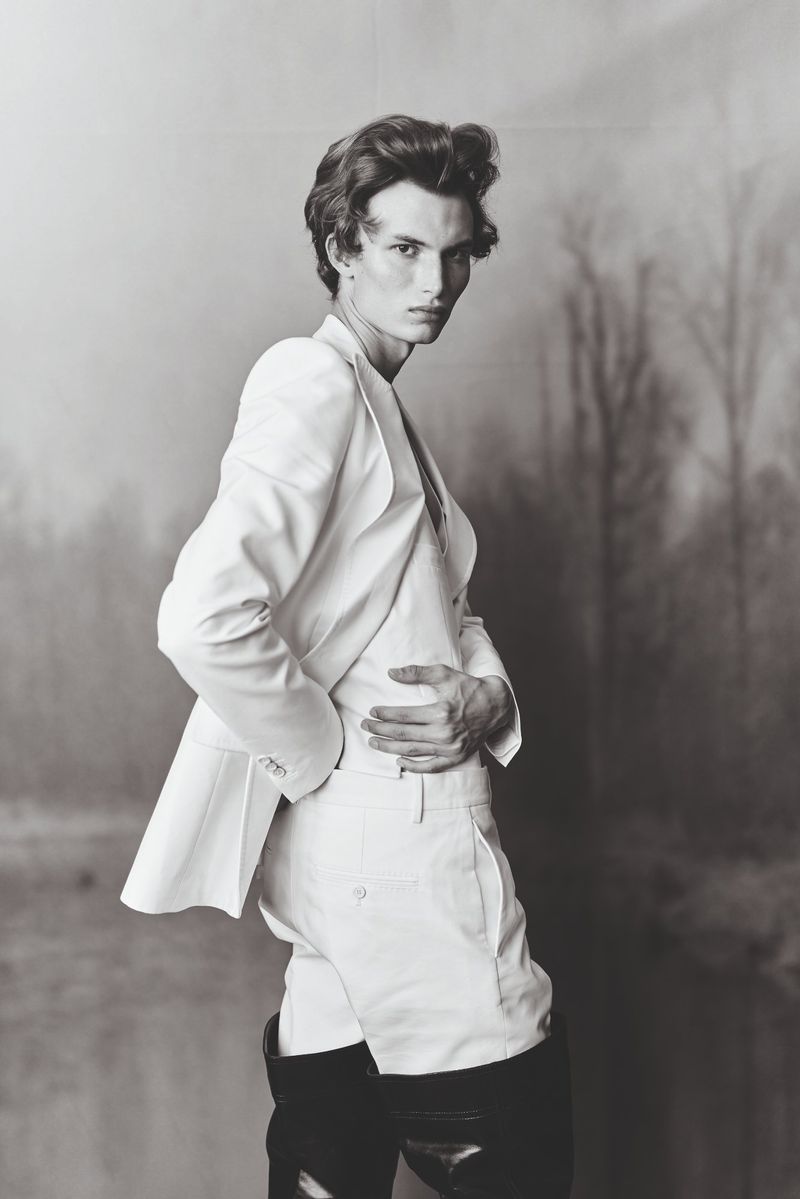 Portrait of a slender male model in a white suit with black boots posing against a blurred black and white nature background.