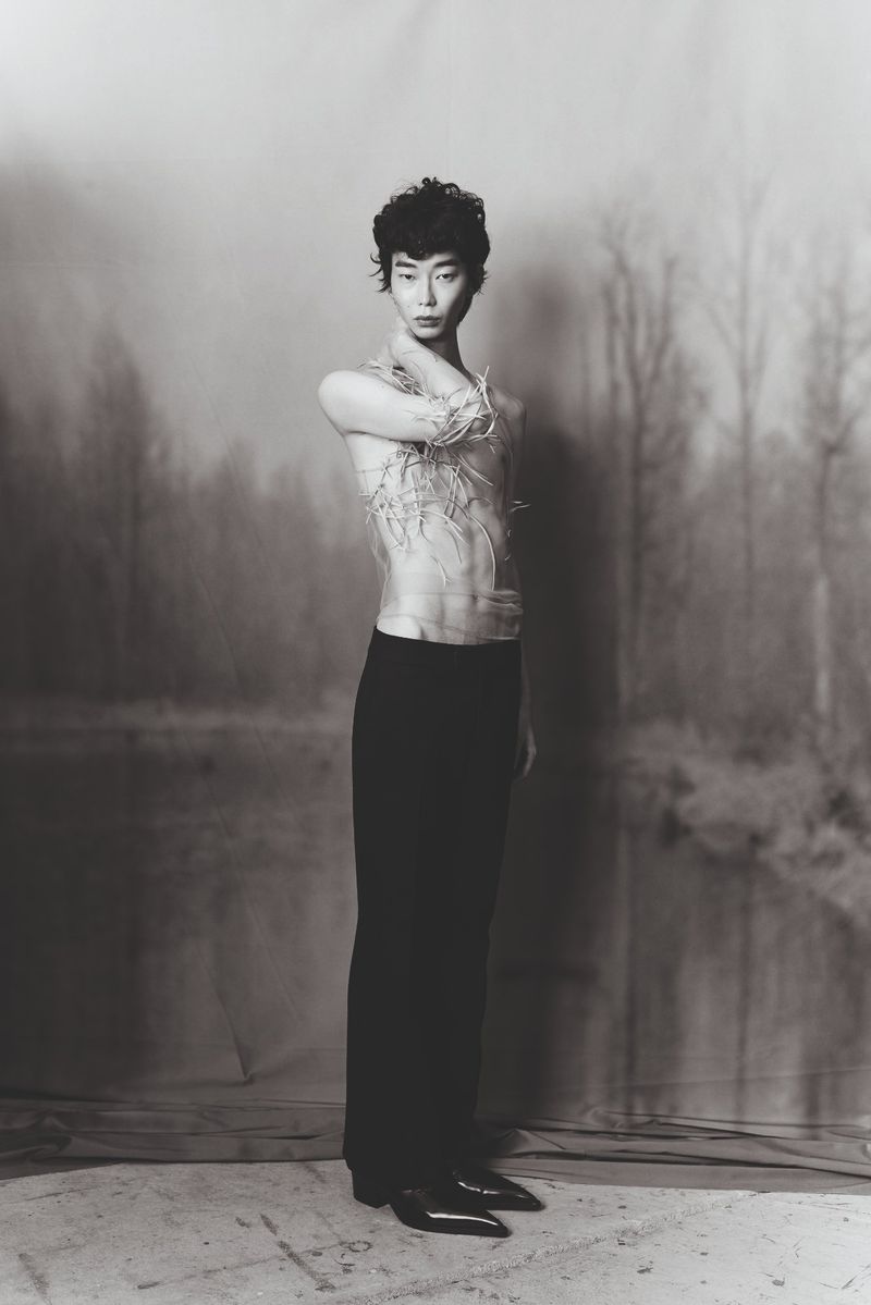 Monochrome portrait of a slender Asian male with curly hair, wearing dark trousers and a sheer textured top, against a muted backdrop.