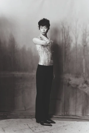 Monochrome portrait of a slender Asian male with curly hair, wearing dark trousers and a sheer textured top, against a muted backdrop.