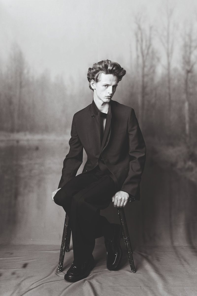 Monochromatic portrait of a young adult male in a suit, seated on a stool against a blurred forest backdrop. Hair is styled, formal attire.