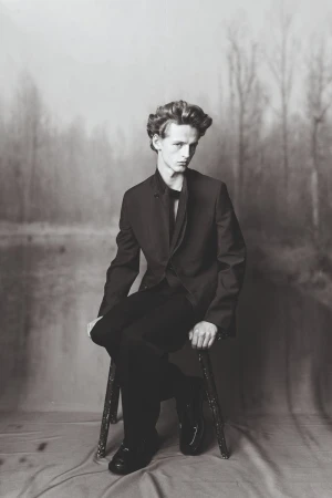 Monochromatic portrait of a young adult male in a suit, seated on a stool against a blurred forest backdrop. Hair is styled, formal attire.
