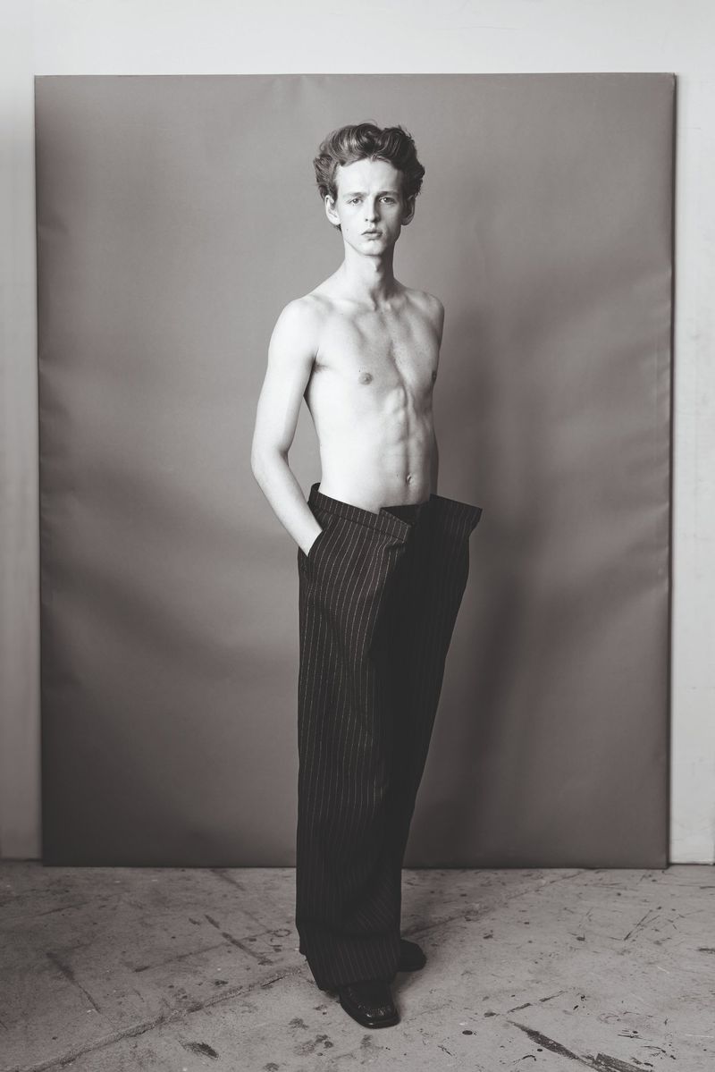 Monochrome portrait of a young adult man with bare chest wearing striped pants standing in a grey photo studio setting.