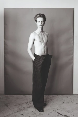 Monochrome portrait of a young adult man with bare chest wearing striped pants standing in a grey photo studio setting.