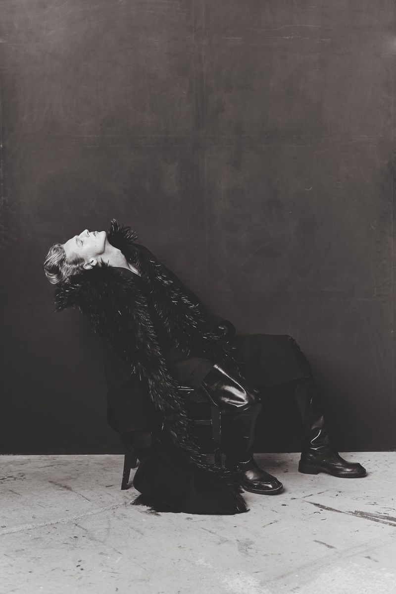 Monochrome fashion shot of a person with an upturned face in a long dark coat, sitting on a chair against a dark backdrop.
