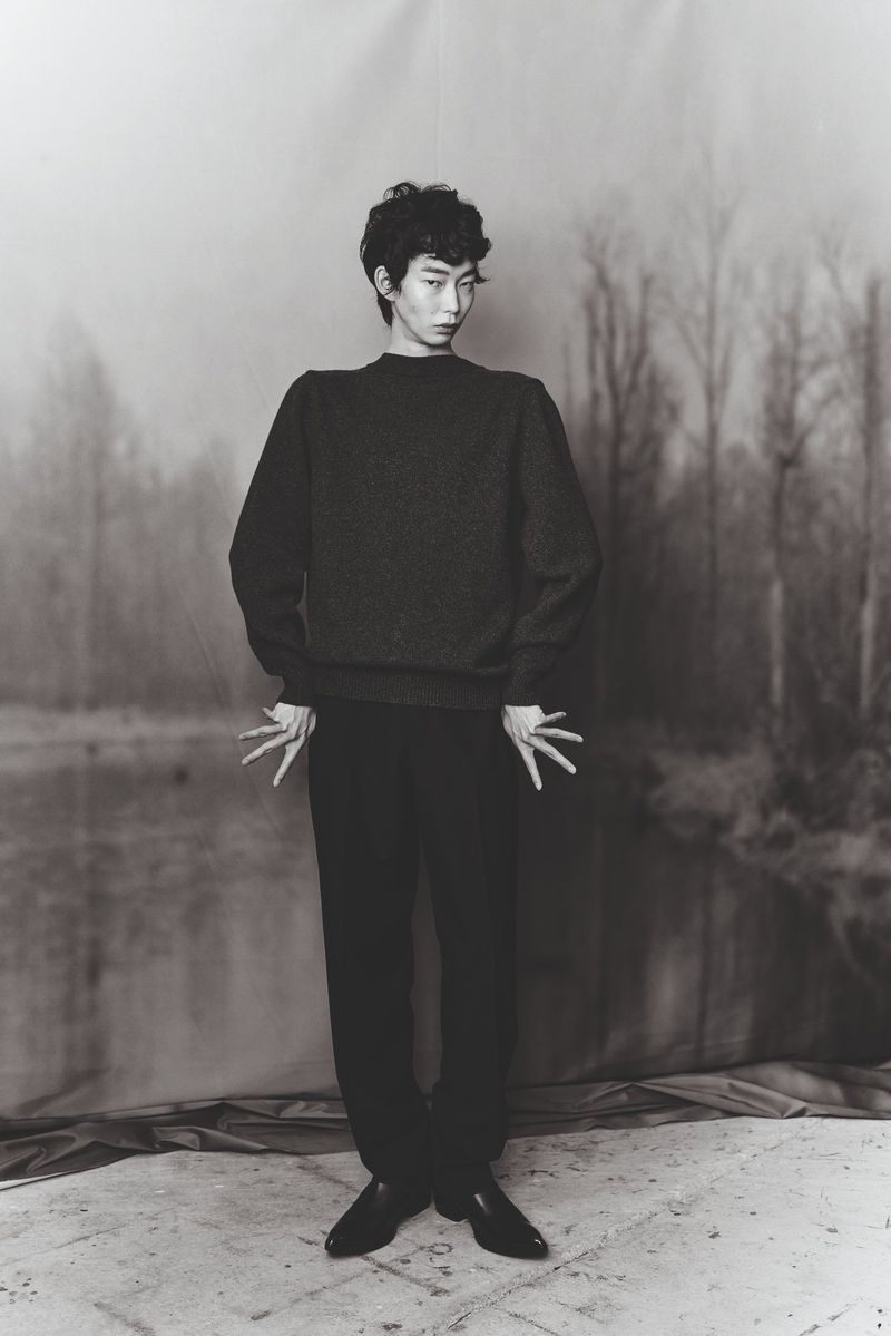 Monochrome portrait of an androgynous person wearing a dark sweater and dark pants posing in front of a blurred landscape backdrop.