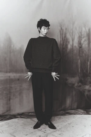 Monochrome portrait of an androgynous person wearing a dark sweater and dark pants posing in front of a blurred landscape backdrop.
