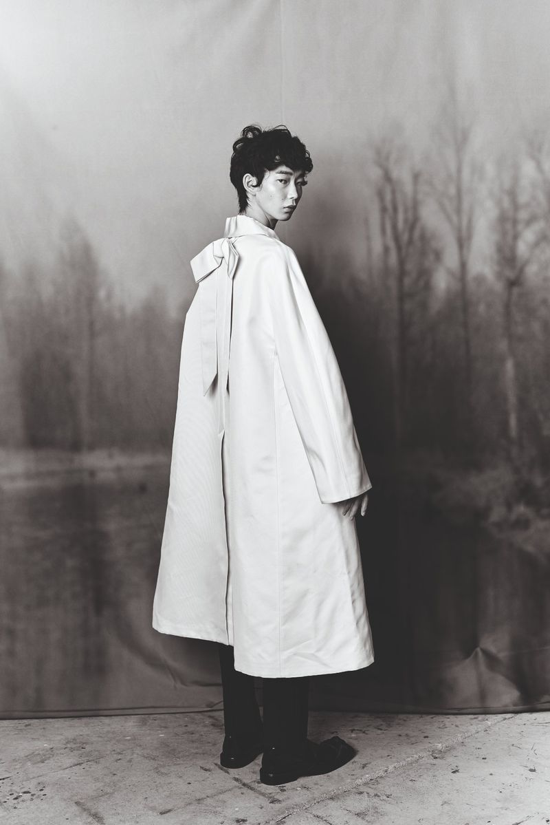 Monochrome fashion shot of a person in a long coat with a bow, posing in front of a backdrop of trees and a body of water.
