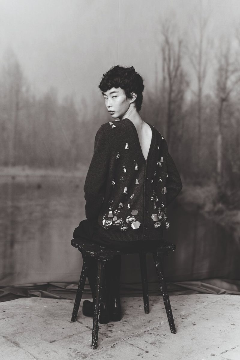 Monochrome image of a seated Asian woman wearing a jeweled sweater with a blurred lake and forest background in soft, neutral tones.