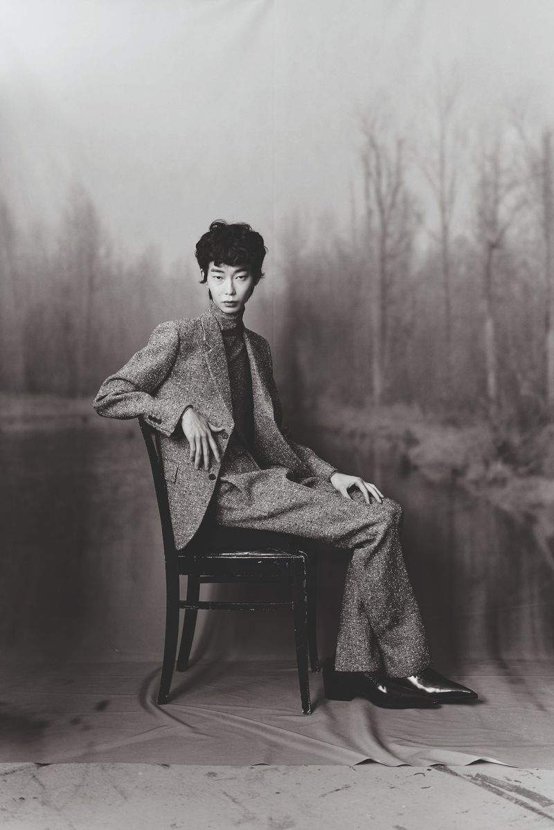 Monochrome studio portrait of a stylish adult in a tweed suit sitting on a chair in front of a blurred forest backdrop. Fashion.