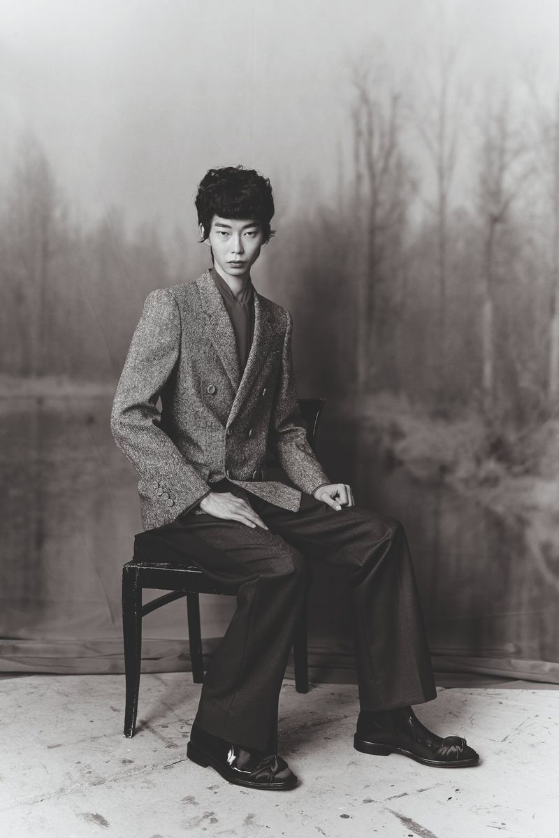 Model Leander Klugmann seated on a wooden chair wearing a textured double-breasted blazer and wide-leg trousers in black and white.
