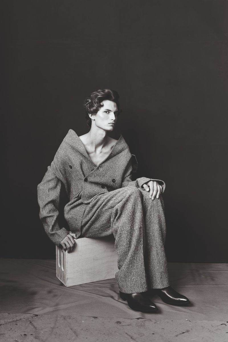 Monochrome portrait of a male model in an oversized tweed suit sitting on a wooden crate against a dark backdrop in a studio setting.