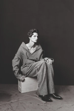 Monochrome portrait of a male model in an oversized tweed suit sitting on a wooden crate against a dark backdrop in a studio setting.