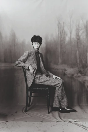 Model seated on a dark wooden chair wearing a textured tweed suit against a painted forest backdrop in black and white.