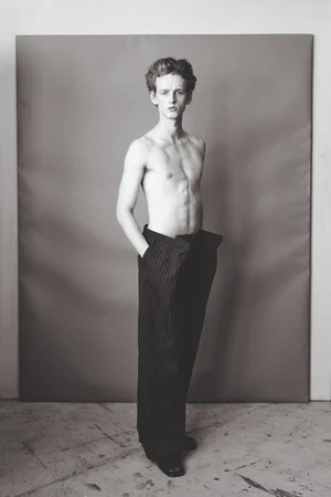 Shirtless male model wearing dark pinstripe trousers against a grey studio seamless backdrop in a black and white portrait.