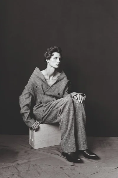 Model Leander Klugmann seated on a wooden crate wearing an oversized off-the-shoulder grey tweed suit against a dark studio backdrop.