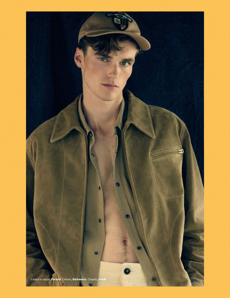 Handsome man in a vintage-style tan jacket and baseball cap poses in a portrait-style fashion shot against a dark backdrop.