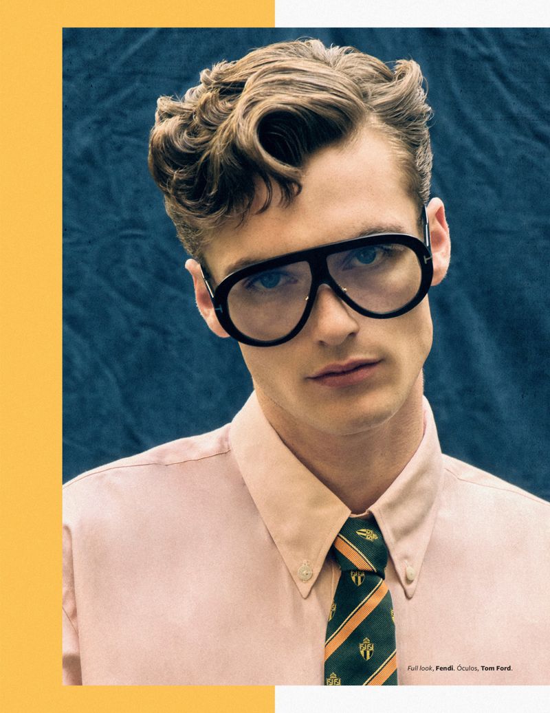 A man with styled hair and large glasses poses wearing a pink shirt and striped tie, in a studio shot with a blue background.