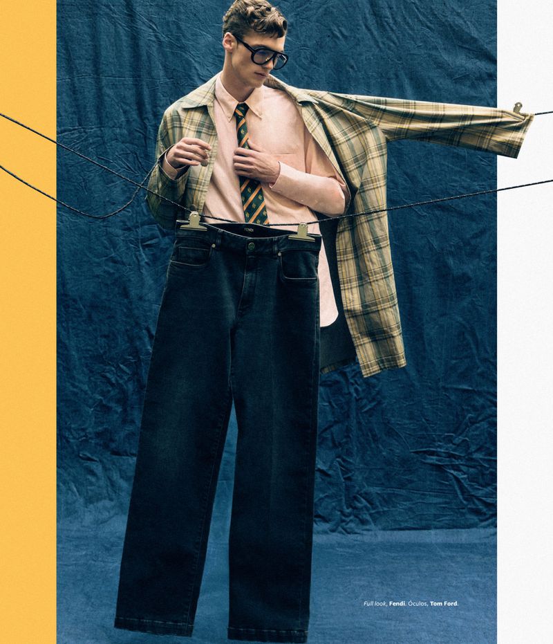 A man poses in fashionable apparel, featuring a checked jacket and dark denim jeans, set against a textured blue background.