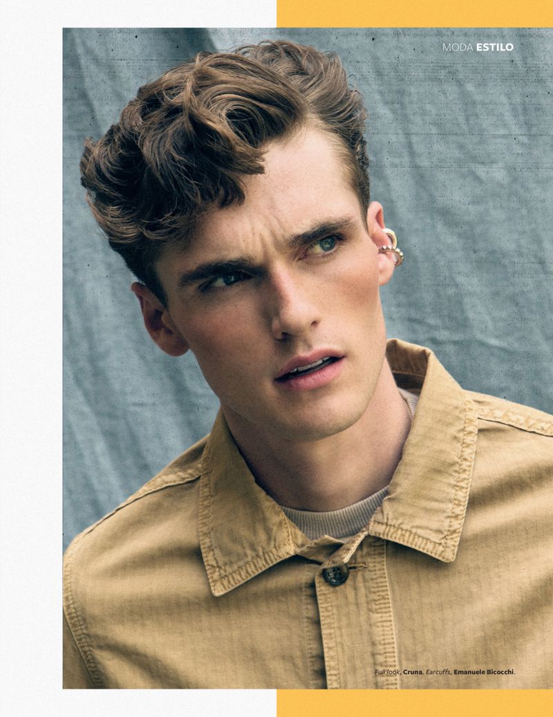Fashion shot of a handsome adult male model with tousled hair wearing a tan button-down shirt in front of a textured blue backdrop.