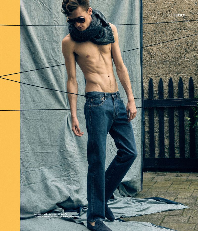 Male fashion model poses shirtless outdoors in jeans and a scarf against a textured backdrop, showcasing a modern, edgy fashion aesthetic.