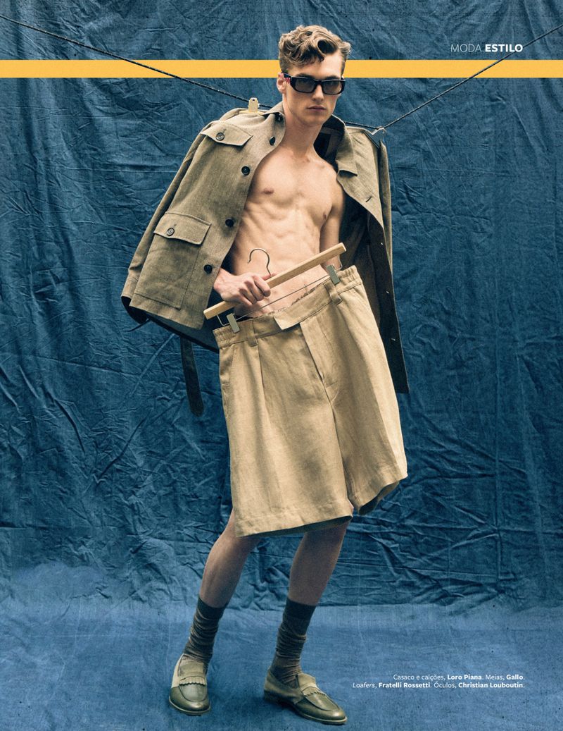 Male fashion model poses in a jacket, shorts, socks, and loafers against a blue backdrop with a yellow stripe in a studio shot.