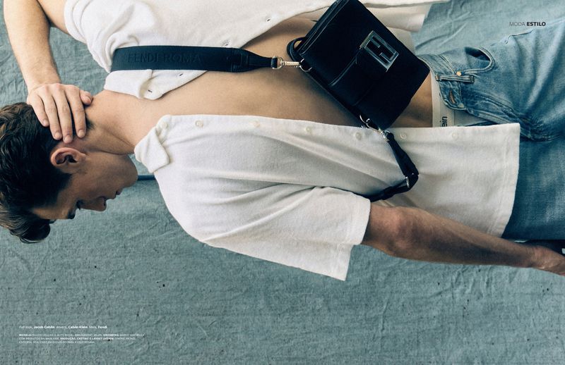 Adult male fashion model lies on a blue surface with a white button-down shirt, light blue denim jeans, and black crossbody purse.