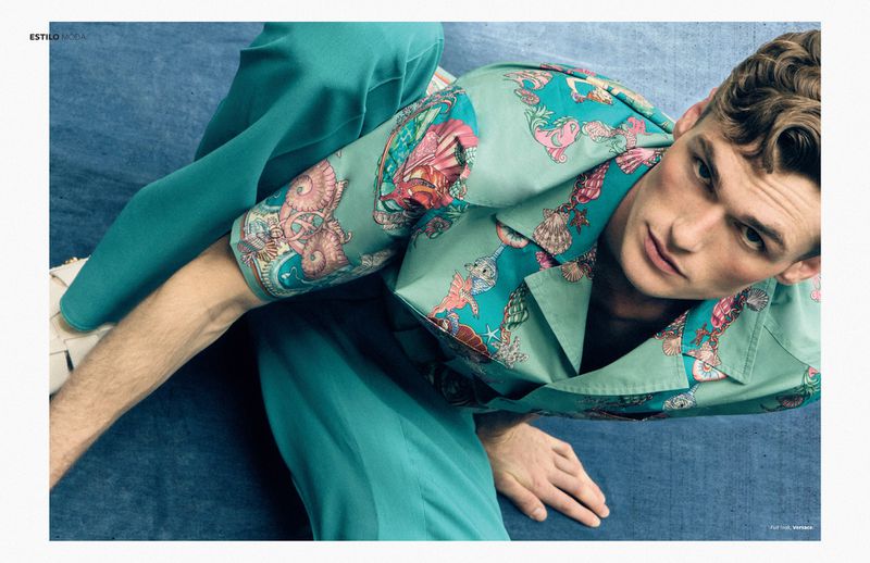 Male fashion model poses in turquoise patterned shirt and pants against a blue textured background in a studio editorial shot.