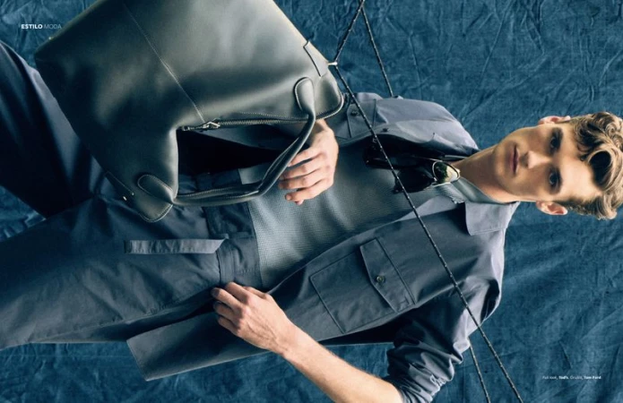 Male model Dillon Collins in a slate grey utility jacket and mesh shirt holding a large leather bag against blue textured fabric.