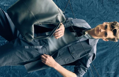 Male model Dillon Collins in a slate grey utility jacket and mesh shirt holding a large leather bag against blue textured fabric.