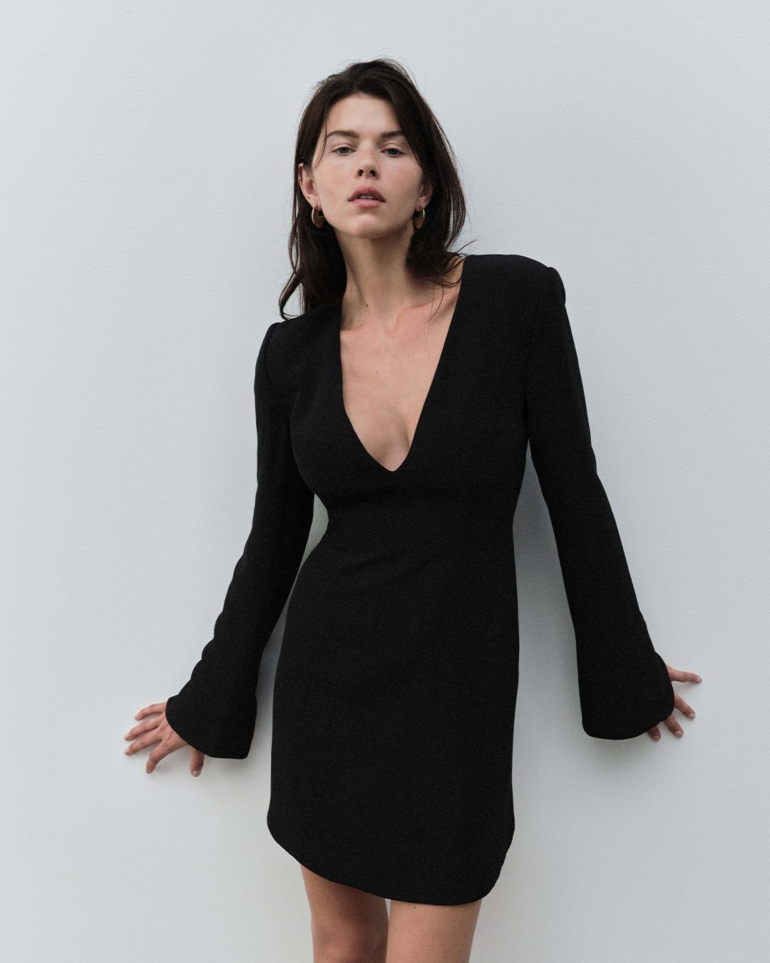 Woman in a long-sleeved black mini dress leaning against a plain light wall in a studio setting