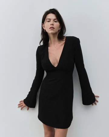 Woman in a long-sleeved black mini dress leaning against a plain light wall in a studio setting
