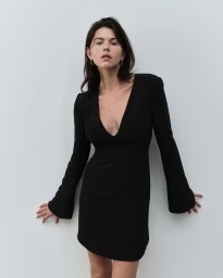 Woman in a long-sleeved black mini dress leaning against a plain light wall in a studio setting