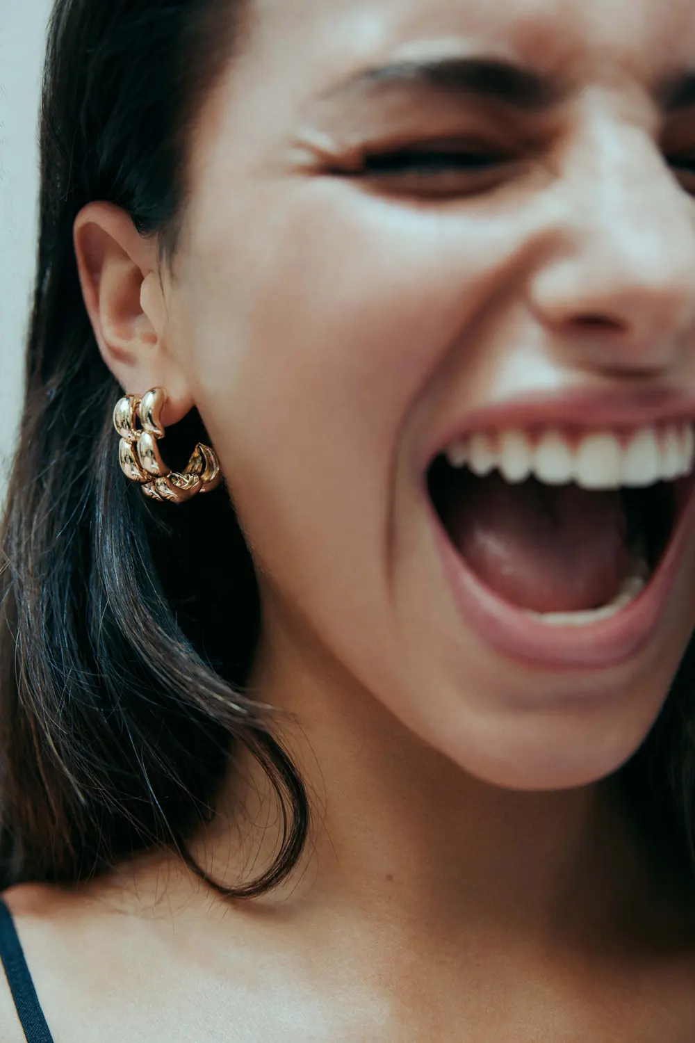Close-up studio shot of a smiling woman with dark hair wearing gold hoop earrings, showcasing her happy expression and jewelry.