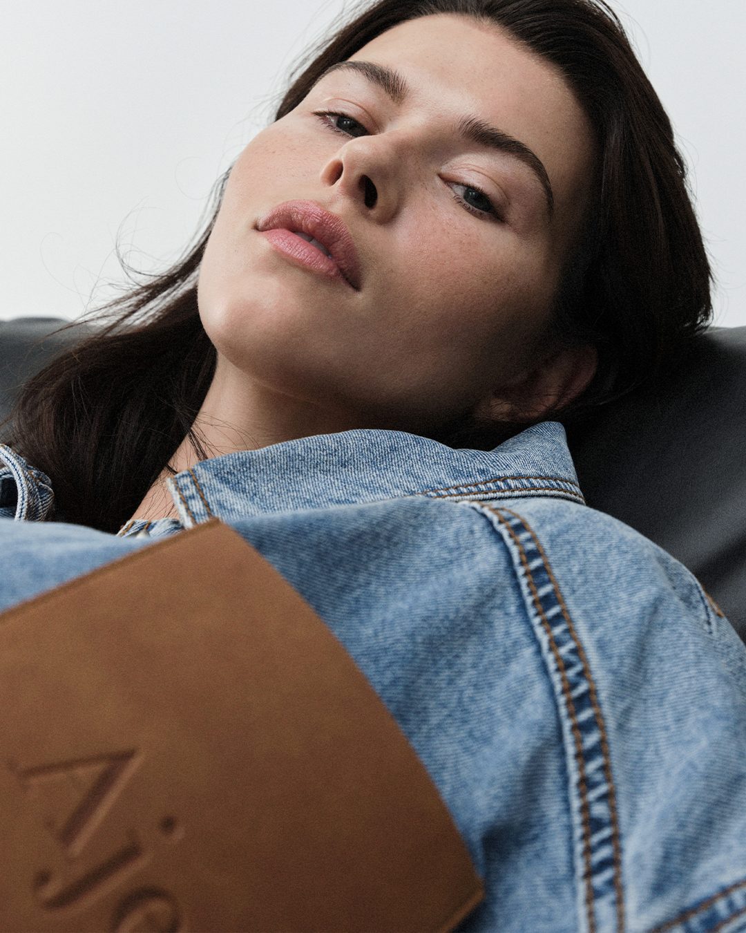 Portrait of a brunette woman in a denim jacket holding a tan leather portfolio in a neutral indoor setting with natural lighting.