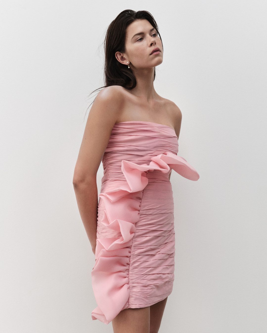 Adult woman posing in a pink strapless mini dress with ruffled detail, set against a plain white backdrop, medium shot.