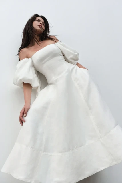 Brunette woman in off-the-shoulder white dress poses indoors against a white wall in a fashion setting, creating an elegant impression.