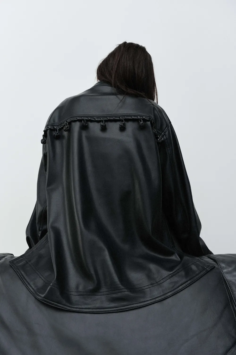 A woman sits with her back to the viewer, wearing a black faux leather cape with decorative tassels on a white background.