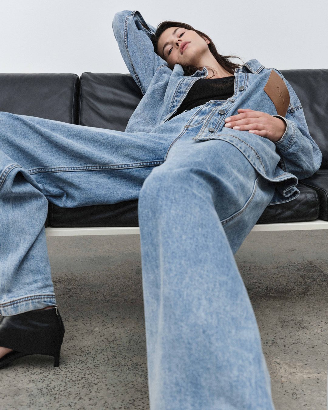 Low-angle shot of a woman in denim clothing reclining on a black leather sofa against a white wall.