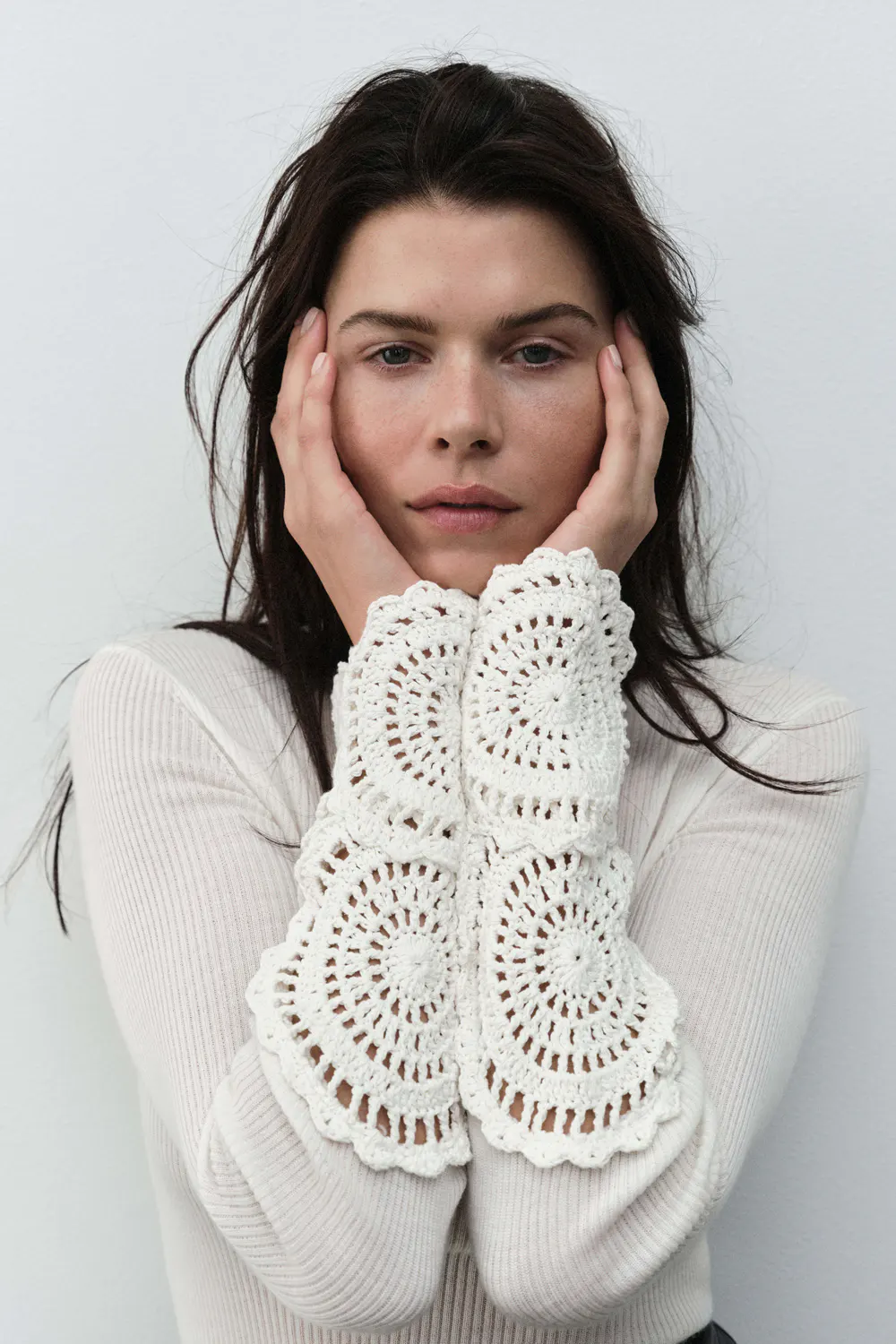 Woman with dark hair poses indoors with her hands against her face while showcasing crochet knit sleeves attached to a long-sleeve shirt.
