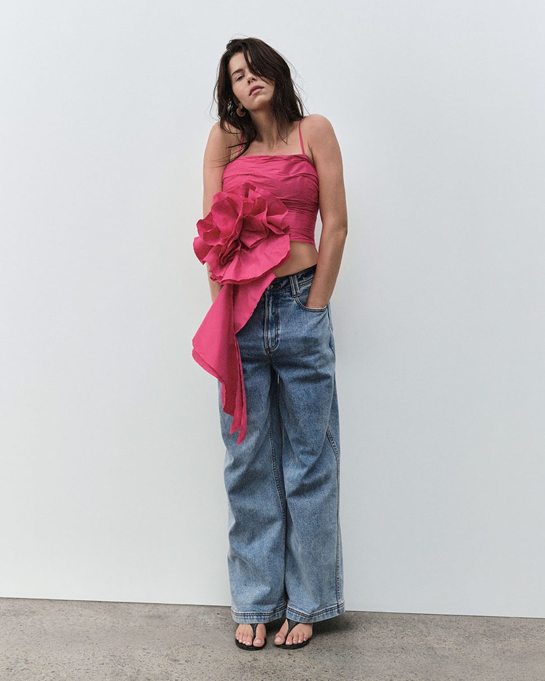 A woman in a pink floral top and jeans stands against a white wall in a fashion shot, posing with a casual, confident attitude.
