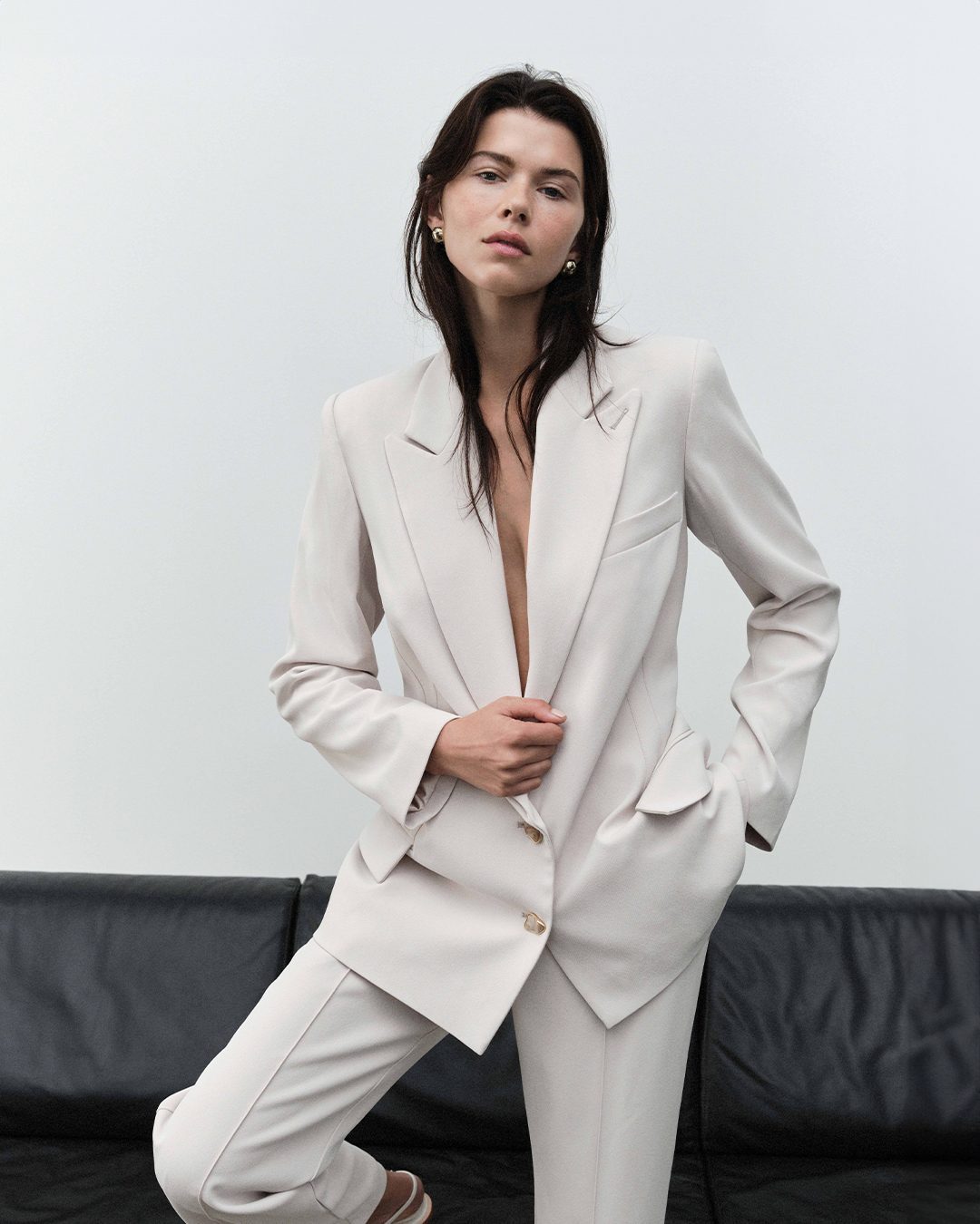 Portrait of a woman in a cream suit, with one leg propped on a black couch, posing in a studio against a white wall, fashion-forward style.
