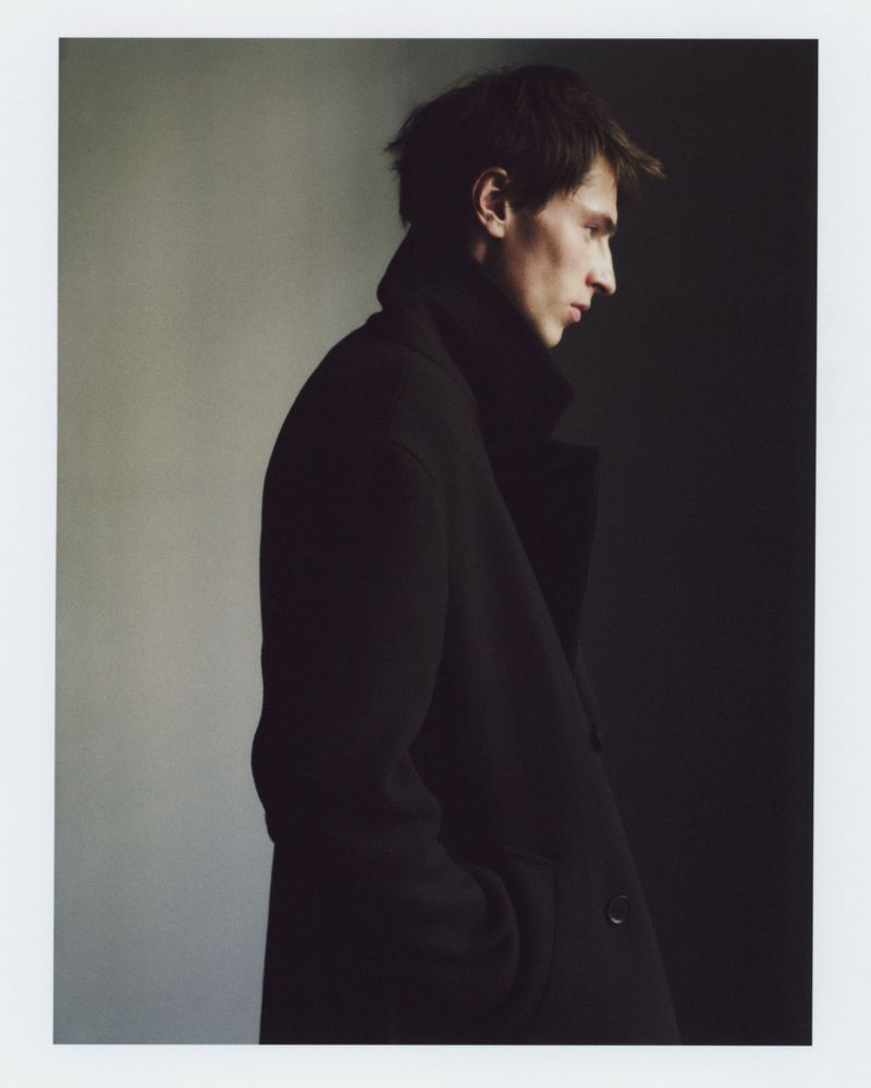 Studio portrait of a young adult man in a dark coat, captured in a profile view against a gradient gray to black background.