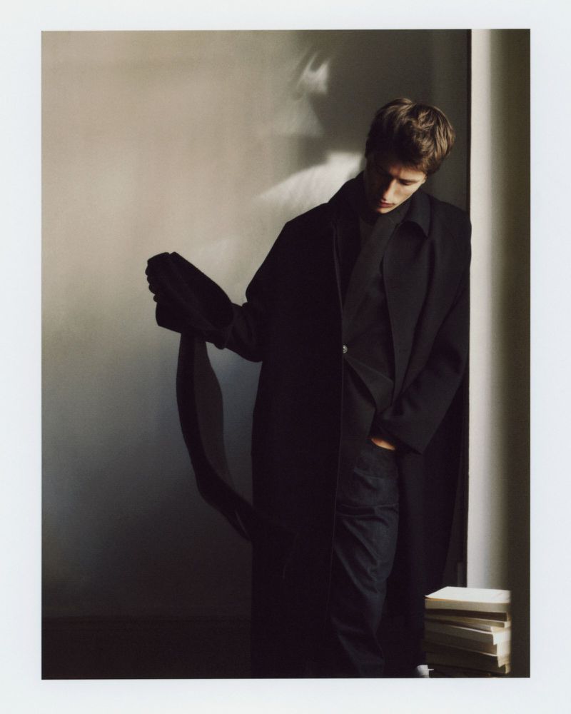 Model wearing an all-black tailored coat and dark trousers standing beside a stack of books against a neutral wall.