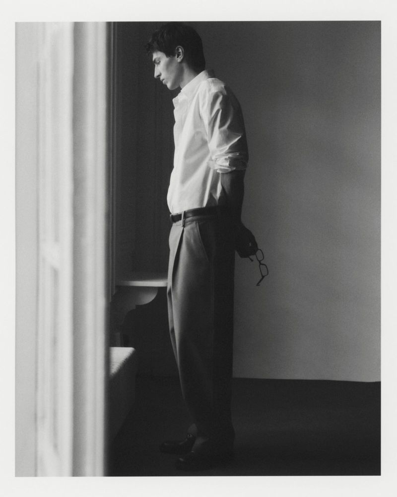 Monochrome portrait of a man in a white shirt and trousers, holding glasses, standing indoors near a window, looking away with thoughtful expression.