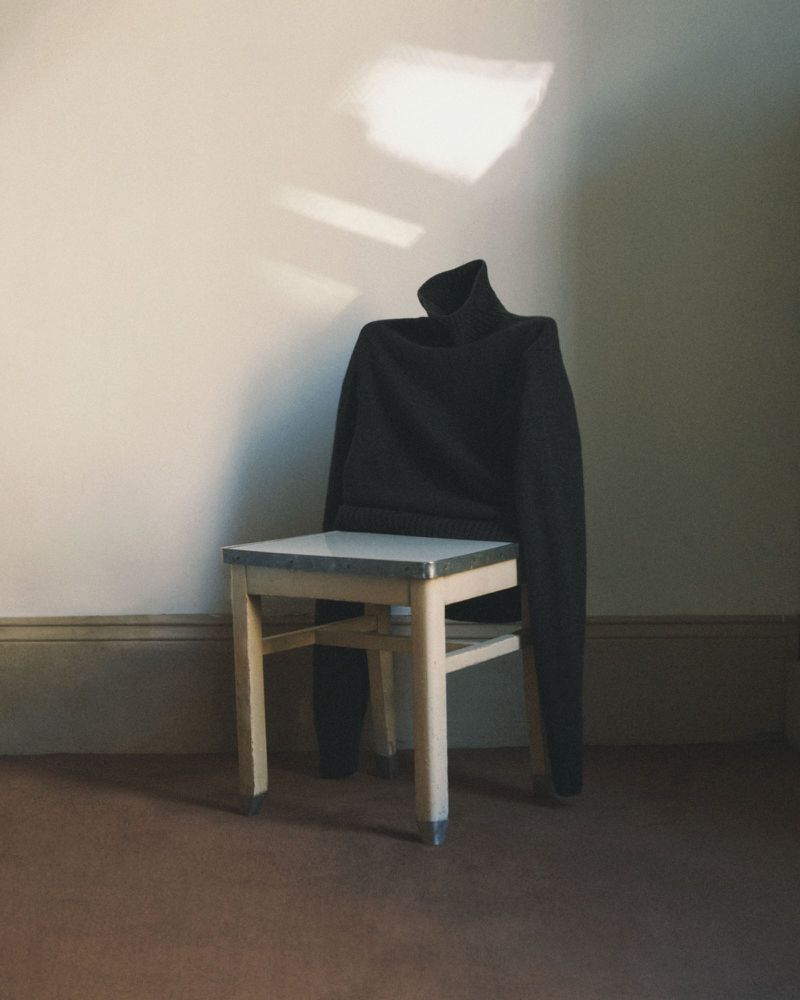 Black turtleneck sweater draped over a wooden chair in a room with natural light creating shadows on the wall.