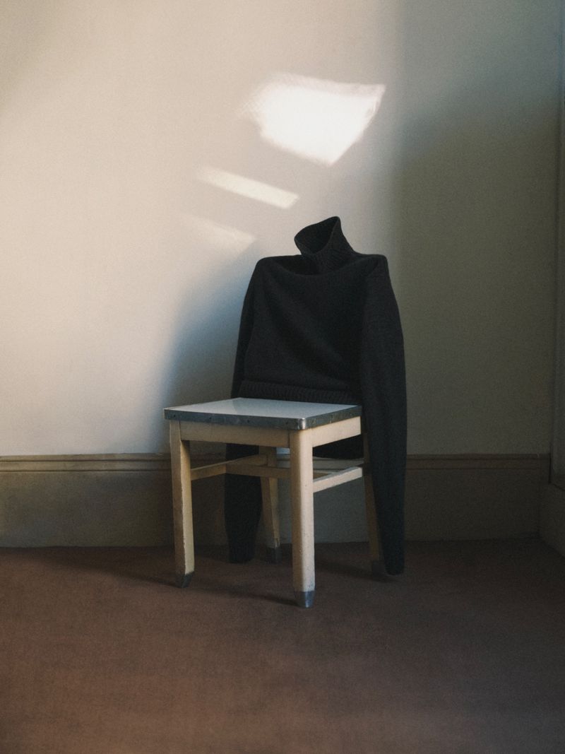 Dark turtleneck sweater draped over a light-colored wooden chair against a neutral wall with natural light creating shadows.