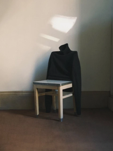 Dark turtleneck sweater draped over a light-colored wooden chair against a neutral wall with natural light creating shadows.