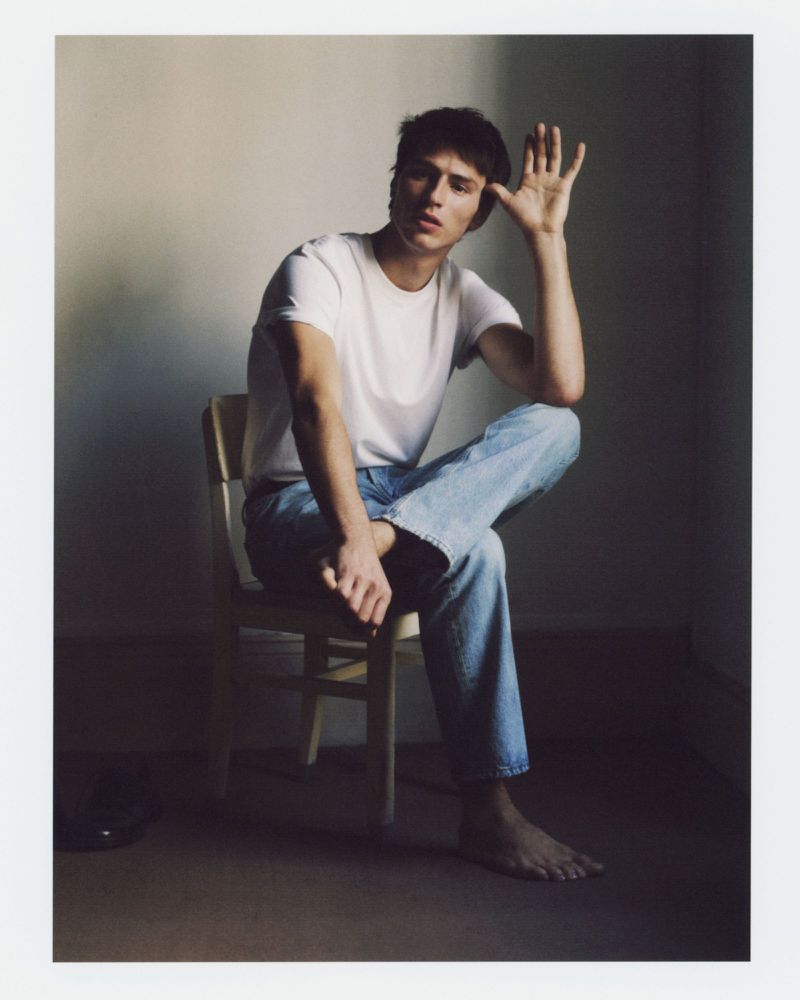 Young man in a white t-shirt and blue jeans sitting on a wooden chair against a plain wall for Zara Origins Fall 2025.